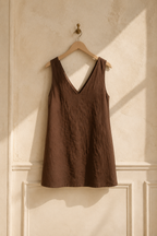 Dark brown linen shift mini dress, hanging on wooden hanger against plaster wall