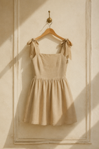 Beige linen a-line mini dress, hanging on wooden hanger against plaster wall