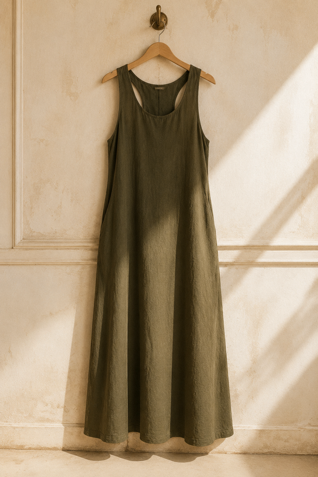 Olive green linen shift maxi dress, hanging on wooden hanger against plaster wall