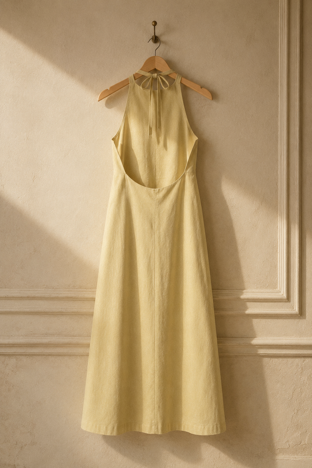 Pale yellow linen shift maxi dress, hanging on wooden hanger against plaster wall