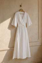 White linen wrap maxi dress, hanging on wooden hanger against plaster wall