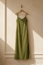 Olive green linen slip maxi dress, hanging on wooden hanger against plaster wall
