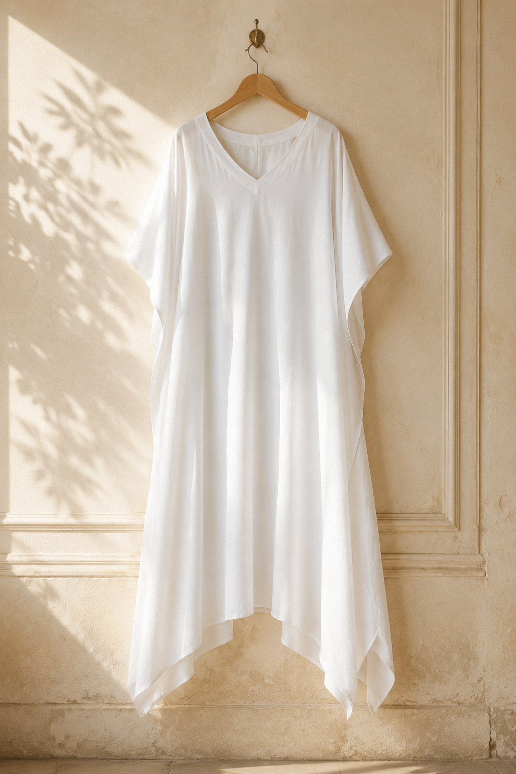 White cotton caftan maxi dress, hanging on wooden hanger against plaster wall