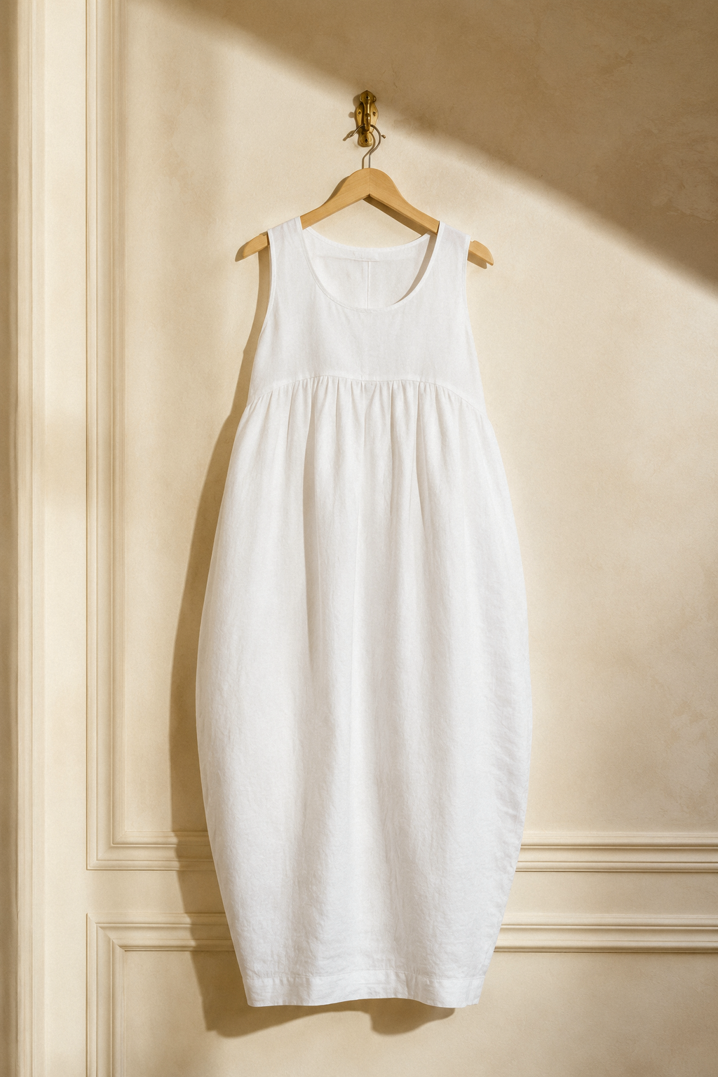 White linen cocoon maxi dress, hanging on wooden hanger against plaster wall