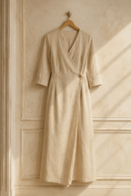 Beige linen wrap maxi dress, hanging on wooden hanger against plaster wall