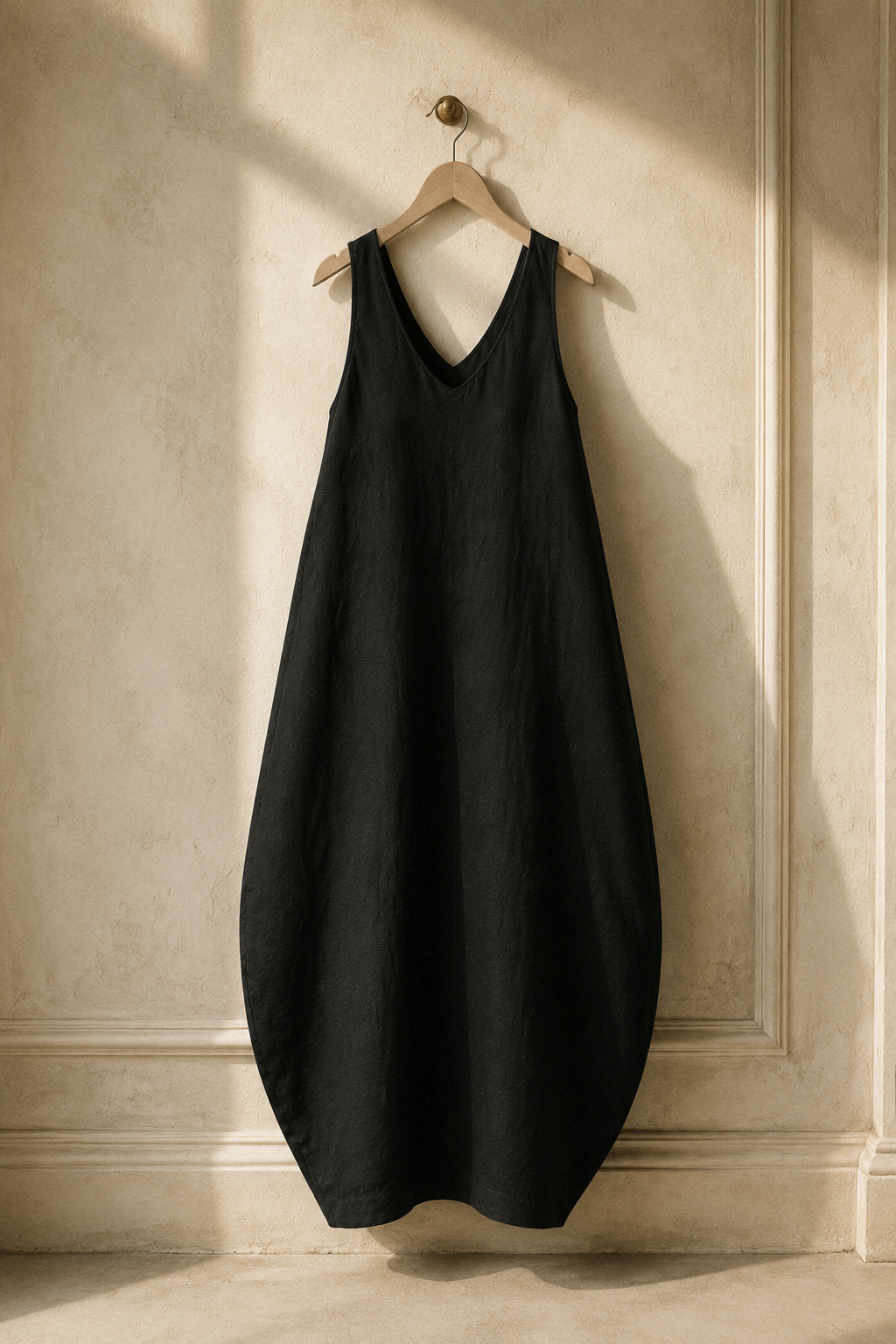 Black linen cocoon maxi dress, hanging on wooden hanger against plaster wall