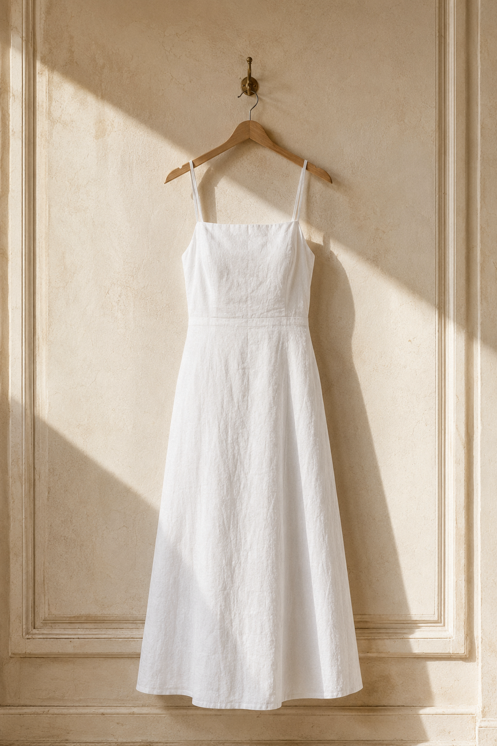 White linen a-line maxi dress, hanging on wooden hanger against plaster wall