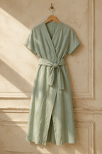 Sage green linen wrap midi dress, hanging on wooden hanger against plaster wall