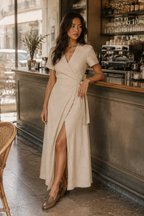 Beige linen wrap midi dress, lifestyle, sun-dappled wall, relaxed posture