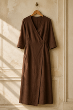 Brown linen wrap midi dress, hanging on wooden hanger against plaster wall