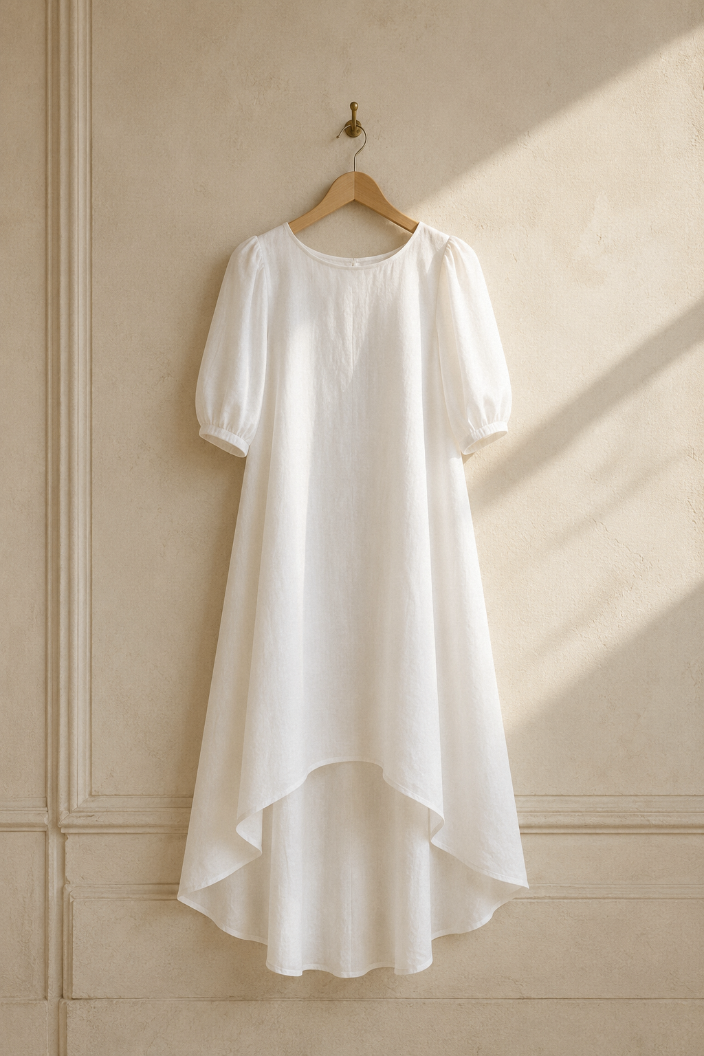 White linen high-low midi dress, hanging on wooden hanger against plaster wall
